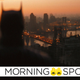 An out of focus Batman in the foreground, faced away from us, looks out over Gotham City at dawn.
